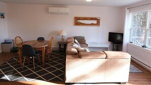 Una sala de estar con un sofá, una mesa de comedor con sillas, un televisor y una ventana con vistas a árboles.