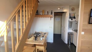 A wooden staircase leading to a dining area with a table set for four, a hanging light fixture, and a kitchen area with utensils hanging above the counter. - Duplex 6 pers at the foot of the slopes Comfort, charm & mountain view Ski in & out (Risoul)