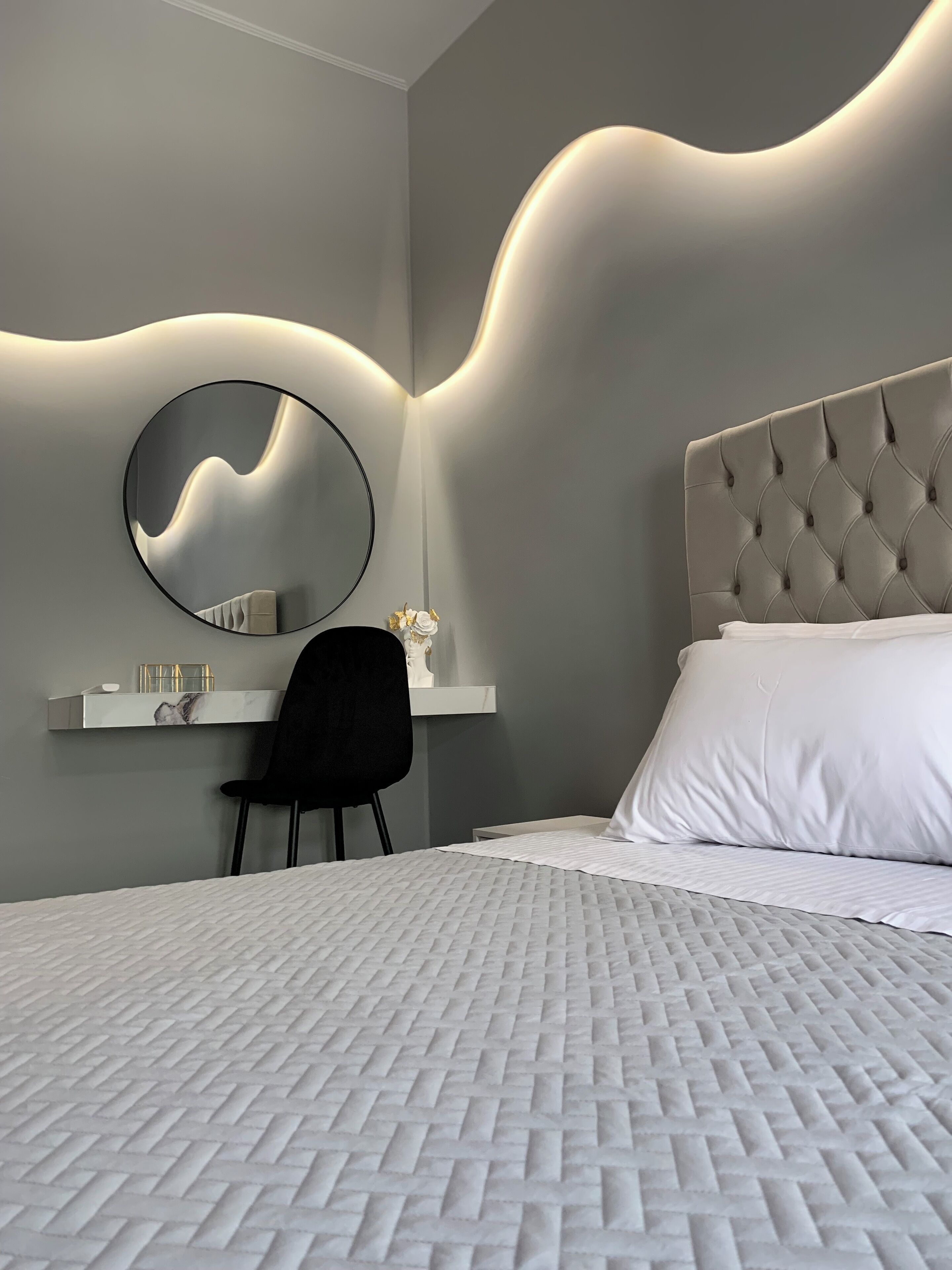 A modern bedroom with a tufted headboard, a round mirror, a desk with a chair, and a neatly made bed.