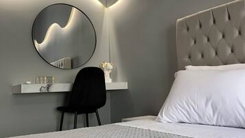 A modern bedroom with a tufted headboard, a round mirror, a desk with a chair, and a neatly made bed.