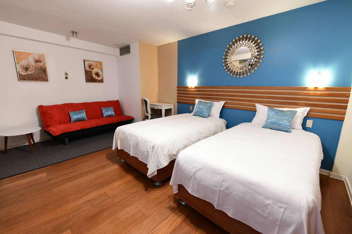 A hotel room with two beds, a red sofa, a small table, and wall art.