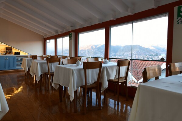 A restaurant with tables set for dining, large windows with a mountain view, and a kitchen area in the background.