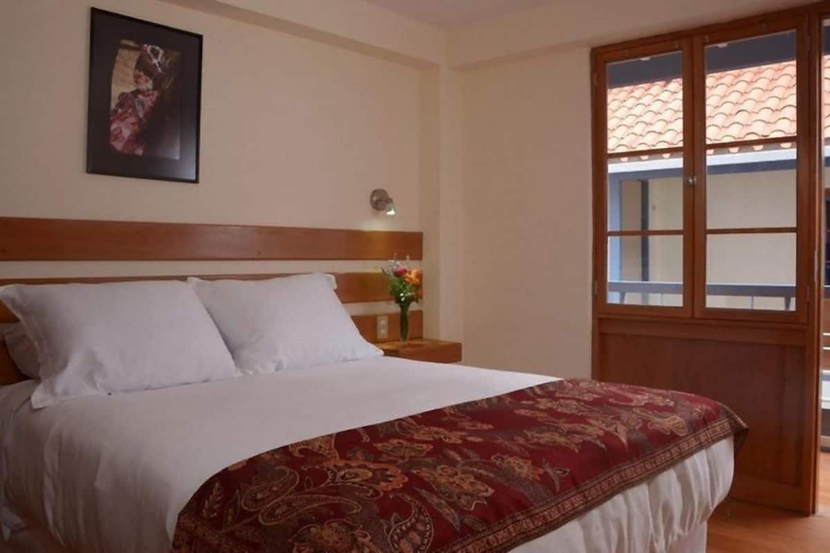A bedroom with a wooden headboard, a large bed with white linens, a red patterned bedspread, a nightstand with a vase of flowers, and a window with a view of a tiled roof.