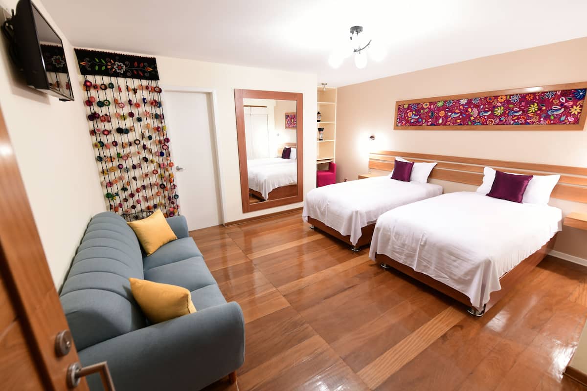 A hotel room with two beds, a sofa, a wooden floor, a TV, a mirror, and a colorful artwork on the wall.