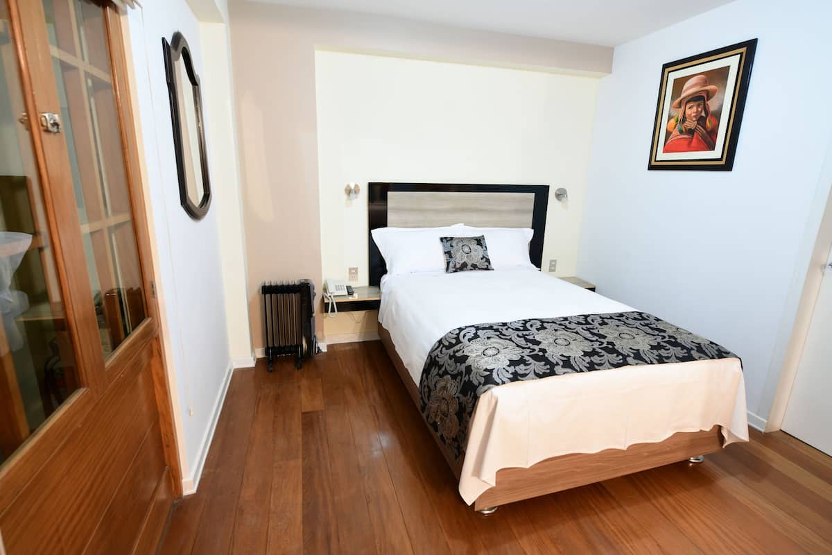 A bedroom with a wooden floor, a bed with a patterned bedspread, a nightstand with a phone, and a framed picture on the wall.