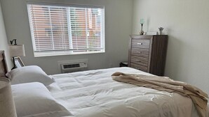 A bedroom with a large bed, a dresser, a window with blinds, and a bedside lamp.