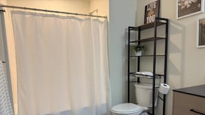 A bathroom with a white toilet, a shower curtain, a white towel on the floor, a black shelf with a plant, and a sign that says "LIVE the life YOU".