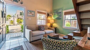 A cozy living room with a sofa, patterned armchair, and a small table. There is a window with a view of the outside, a lamp on a side table, and a decorative wall piece. - Margaretaville (Key West)