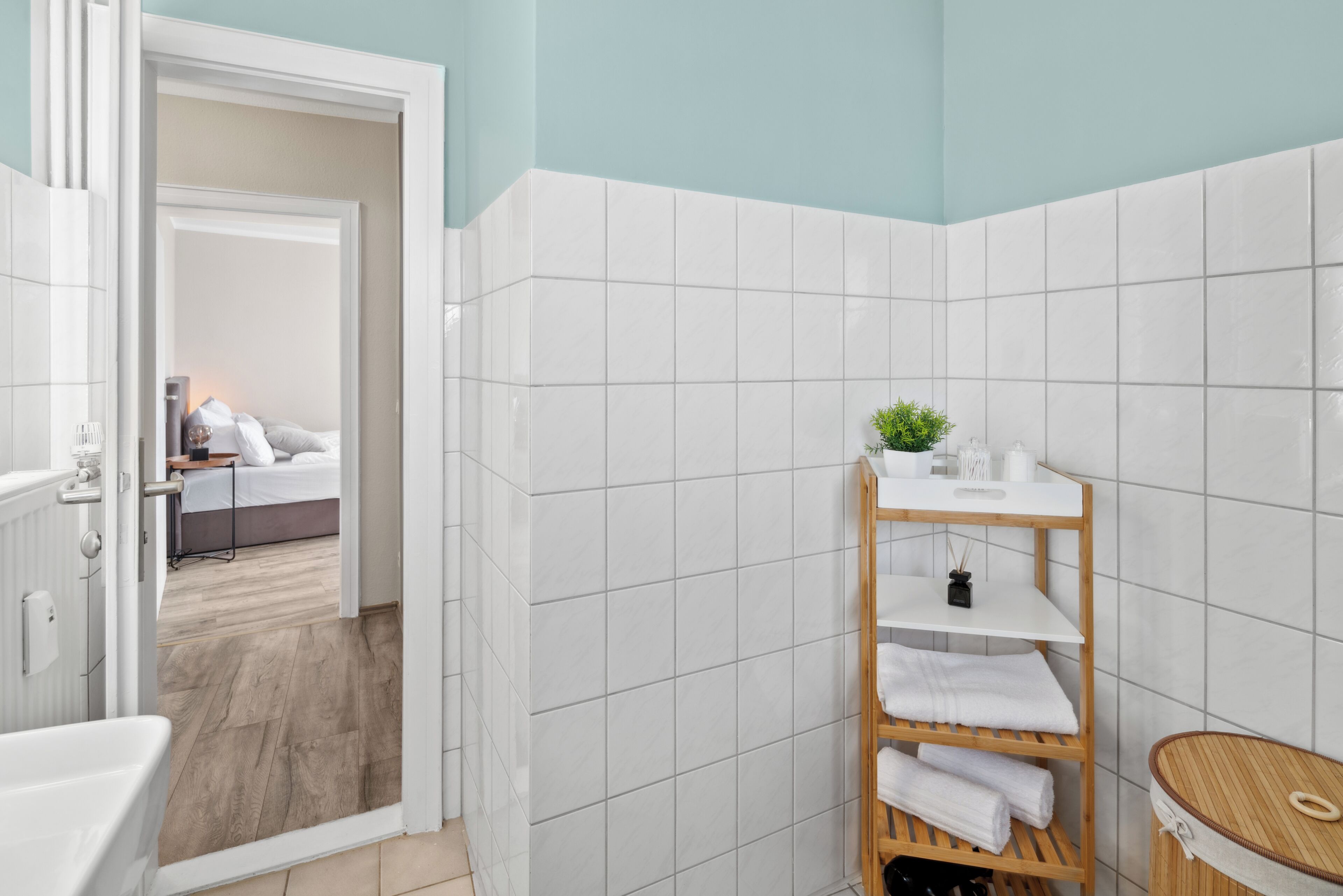 Ein Badezimmer mit weißen Fliesen, einem hölzernen Ablagebrett mit Handtüchern, einer kleinen Pflanze und einem Holzschemel.