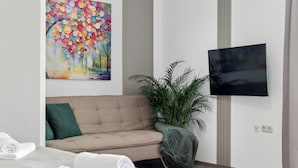 Camera d'albergo moderna con un divano color beige, una grande pianta in vaso, un televisore a schermo piatto montato a parete e un letto rifatto con asciugamani bianchi arrotolati.