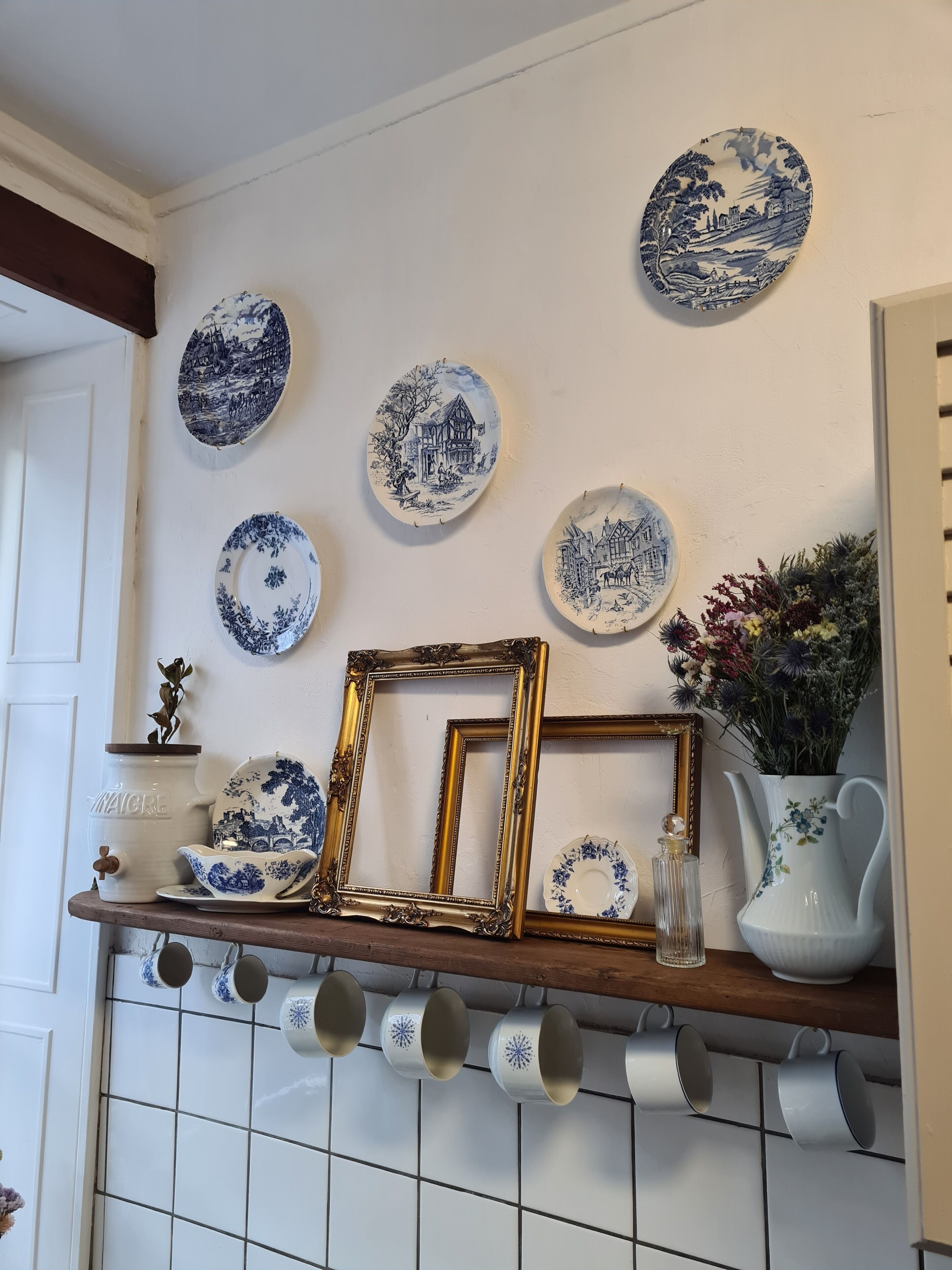 Un mur orné de différents modèles d’assiettes décoratives et une étagère en bois sur laquelle sont posés des photos encadrées, un pichet et un vase rempli de fleurs.