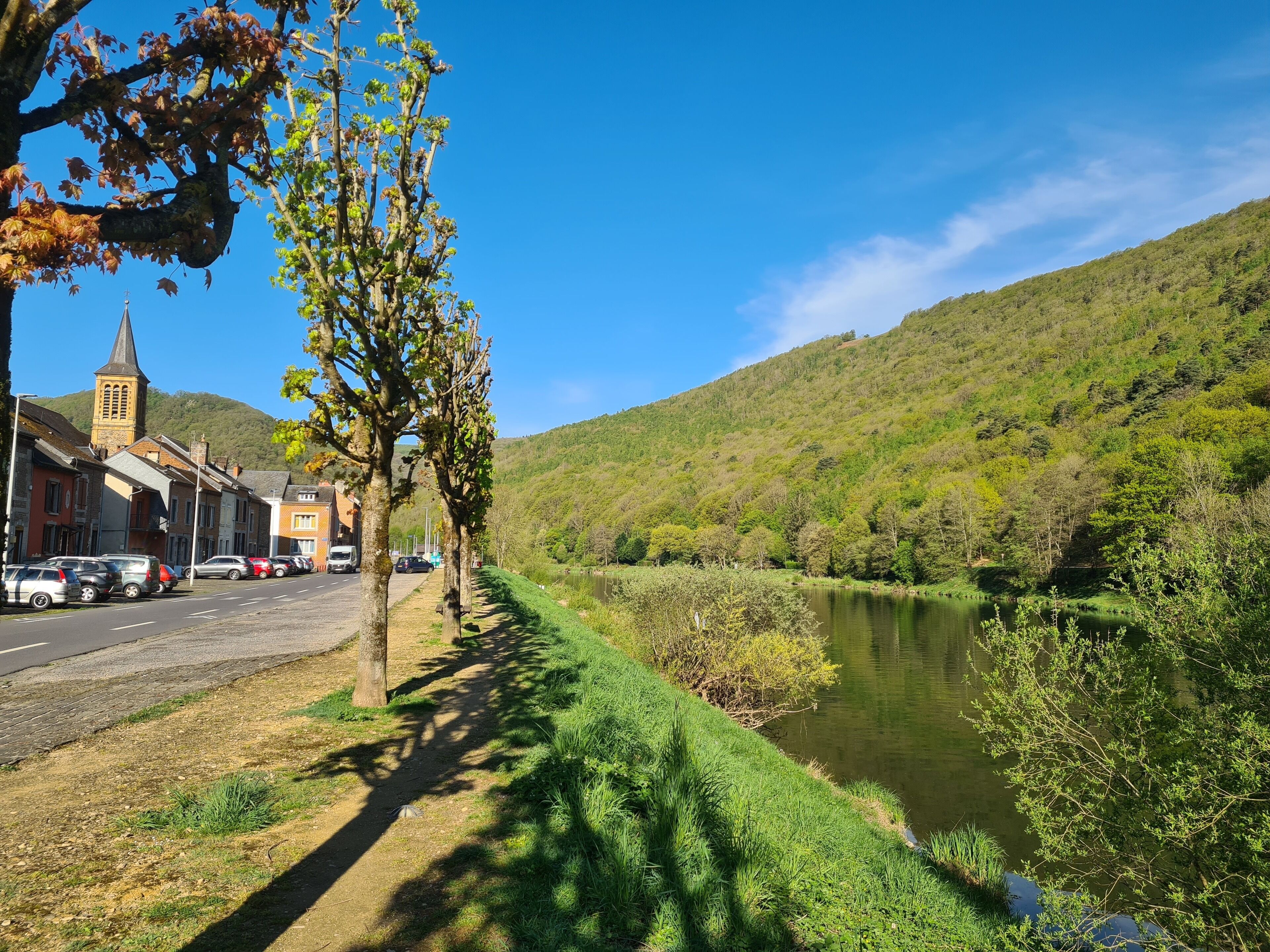 Une route longeant la rivière, bordée de voitures garées, une église et une colline couverte d’une végétation luxuriante.