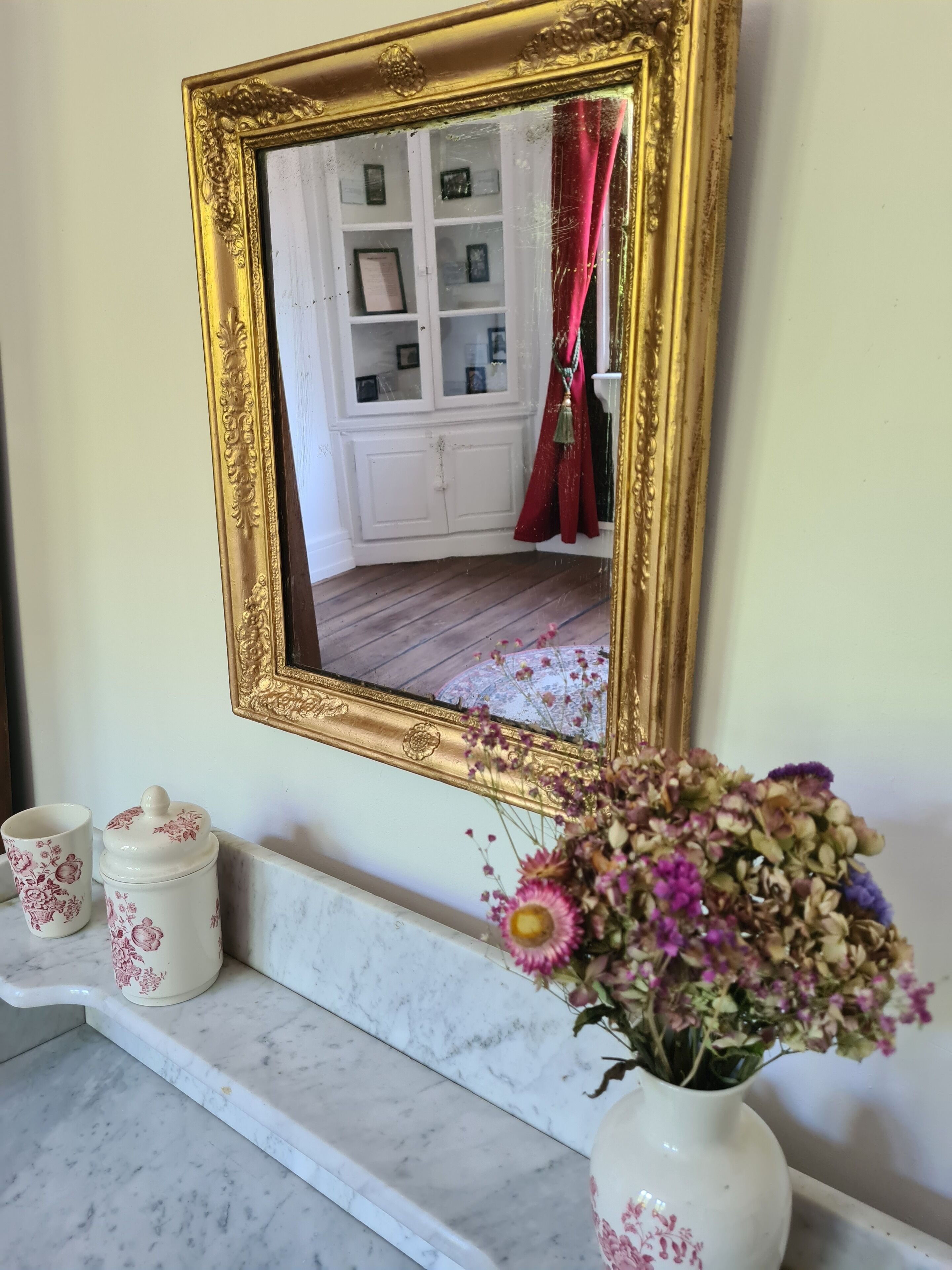 A bathroom with a gold-framed mirror, a marble countertop, a vase with flowers, and a cabinet with framed pictures.