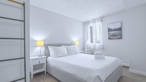 A modern, minimalist bedroom with a large bed, two bedside tables, and a window with curtains. - Cozy & Spacious 2BR at Stampede w/ Parking, AC (Calgary)