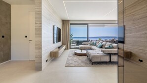 Ein modernes Wohnzimmer mit einer großen Couch, einem Couchtisch und einem Fernseher. Große Fenster bieten Blick auf den Ozean.
