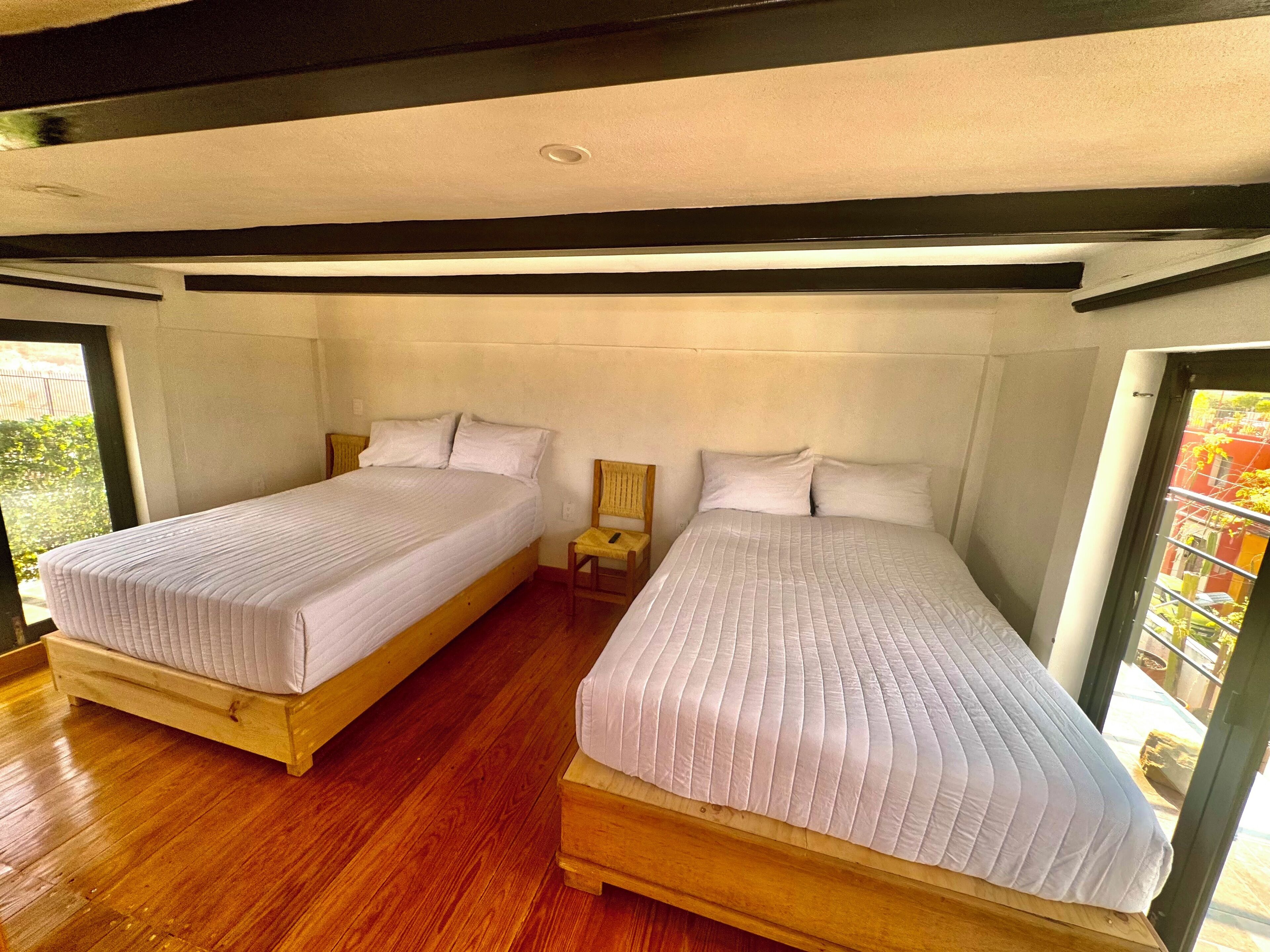 A room with two beds, wooden flooring, and a window providing a view of greenery outside.