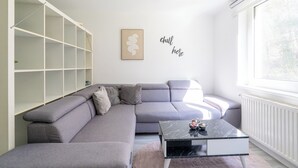 Un salon moderne avec un canapé d’angle gris, une table basse en marbre noir et un radiateur blanc. Un meuble à étagères blanc se trouve à gauche et une fenêtre à droite.
