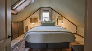 Een slaapkamer met een schuine kap, een bed met hoofdeinde, een nachtkastje met lamp, een raam met gordijnen en een deur.