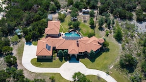 An aerial view of a large, multi-story house with a swimming pool, surrounded by trees and a landscaped driveway.