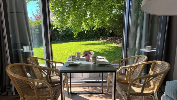 Un coin repas avec des chaises en osier, une table et une vue sur une pelouse verdoyante, visible à travers de grandes fenêtres.