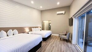 Une chambre d’hôtel avec deux lits, un coin salon avec un fauteuil, une grande fenêtre et un appareil de climatisation.