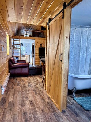 L’intérieur chaleureux d’un chalet avec un plancher en bois, un canapé, un téléviseur et une porte coulissante donnant accès à une salle de bain.