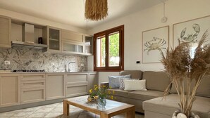 A modern kitchen with a window, a wooden table with flowers, a sofa, and a vase with dried plants.