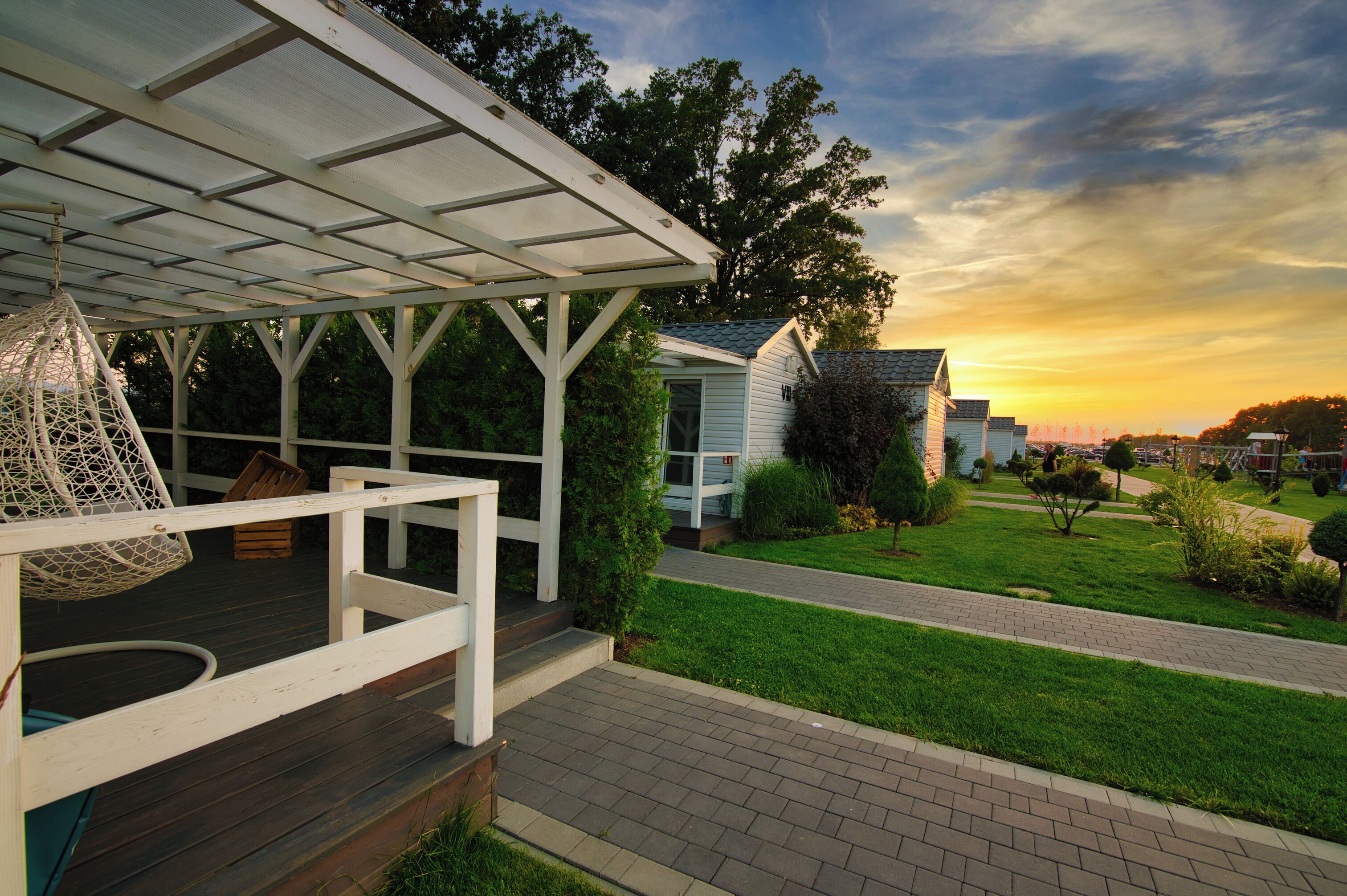 A covered outdoor deck with a hanging chair, a small house with a sloped roof, and a well-maintained lawn with a paved walkway.