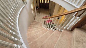 Un magnifique escalier avec des balustrades blanches et une rampe en bois, menant à un palier d’où l’on peut admirer une pièce dotée d’une grande fenêtre et d’une plante en pot.