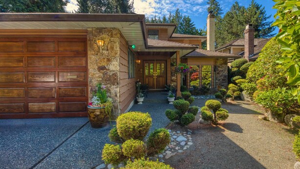 Une maison moderne avec une façade en pierre, un garage en bois et un jardin bien entretenu agrémenté de divers arbustes et de plantes en pot.