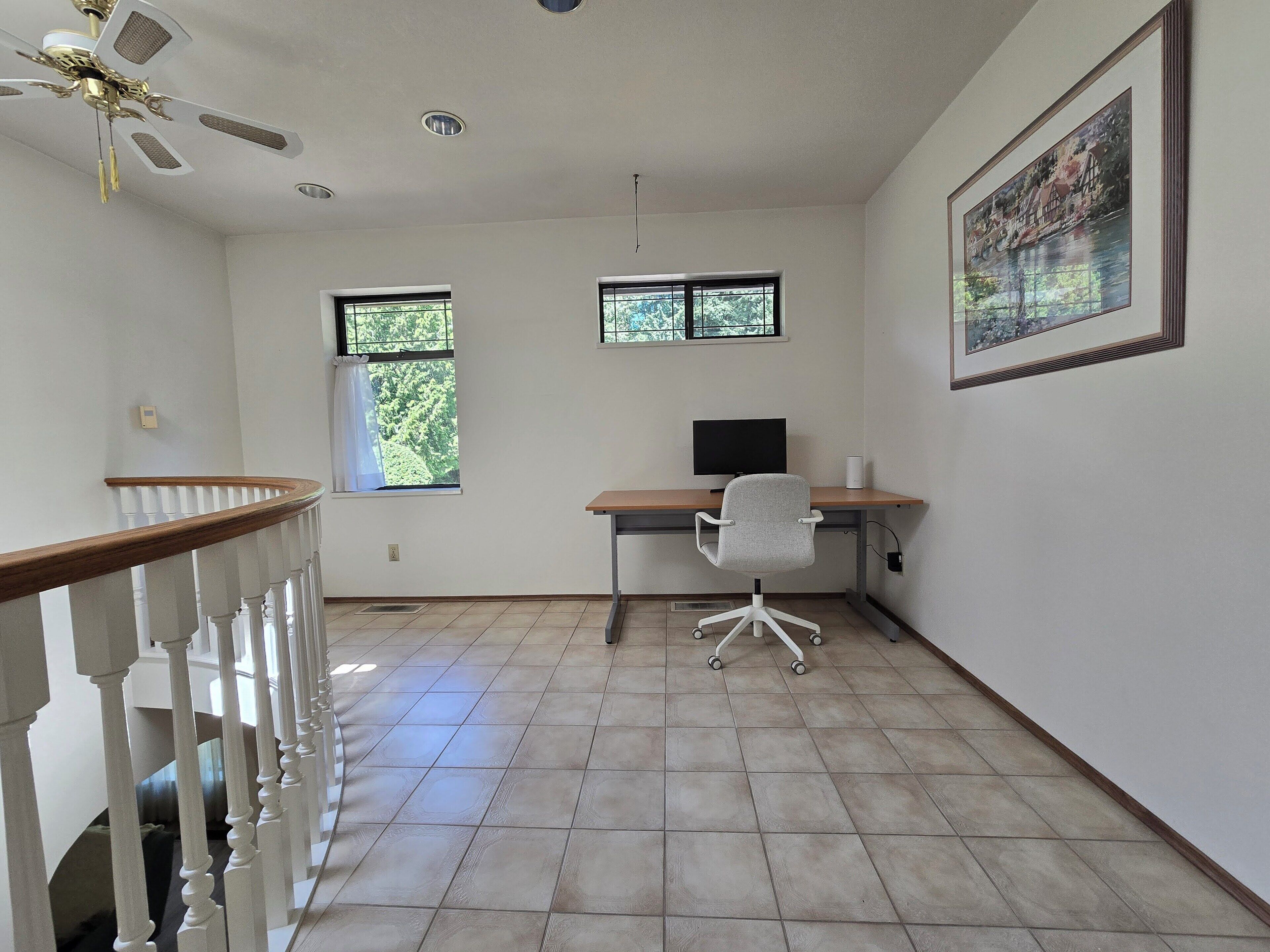 A room with a desk, chair, and a ceiling fan. There are two windows with curtains, a painting on the wall, and a staircase with a wooden railing.