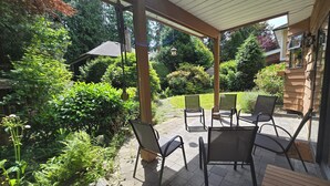 Un patio couvert avec des sièges d’extérieur, entouré d’une végétation luxuriante et offrant une vue sur une maison.