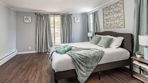 Un dormitorio con una cama grande, cortinas grises, una obra de arte colgada en la pared y una mesita de noche con una lámpara.
