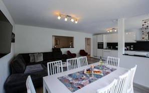 A modern living room with a dark-colored sofa, white chairs, and a dining table set with a colorful table runner. A kitchen with white cabinets and a black countertop is visible in the background.