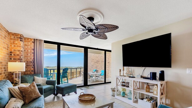 A modern living room with a large TV, comfortable seating, a ceiling fan, and a view to the outside.