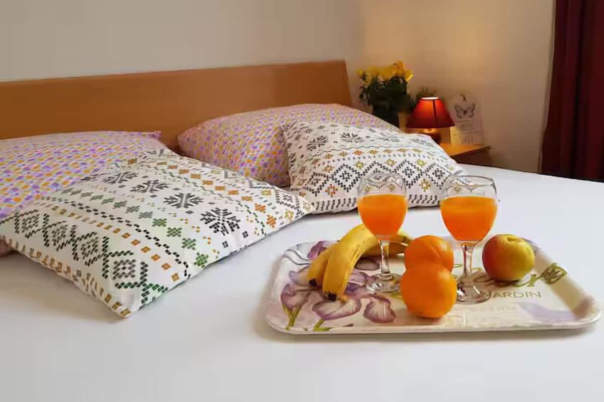 Een netjes opgemaakt bed met gestreepte kussens, een dienblad met fruit en sapglazen, en een nachtkastje met een lamp en bloemen.