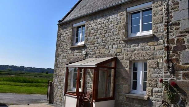 A stone house with a glass door leading to a room, a window, and a view of a green field. - Gite des Bords de rance (Pleudihen-sur-Rance)