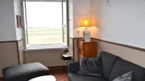 A room with a grey sofa, a small round table, a window with a view of a field, and a lamp on a wooden cabinet. - Gite des Bords de rance (Pleudihen-sur-Rance)
