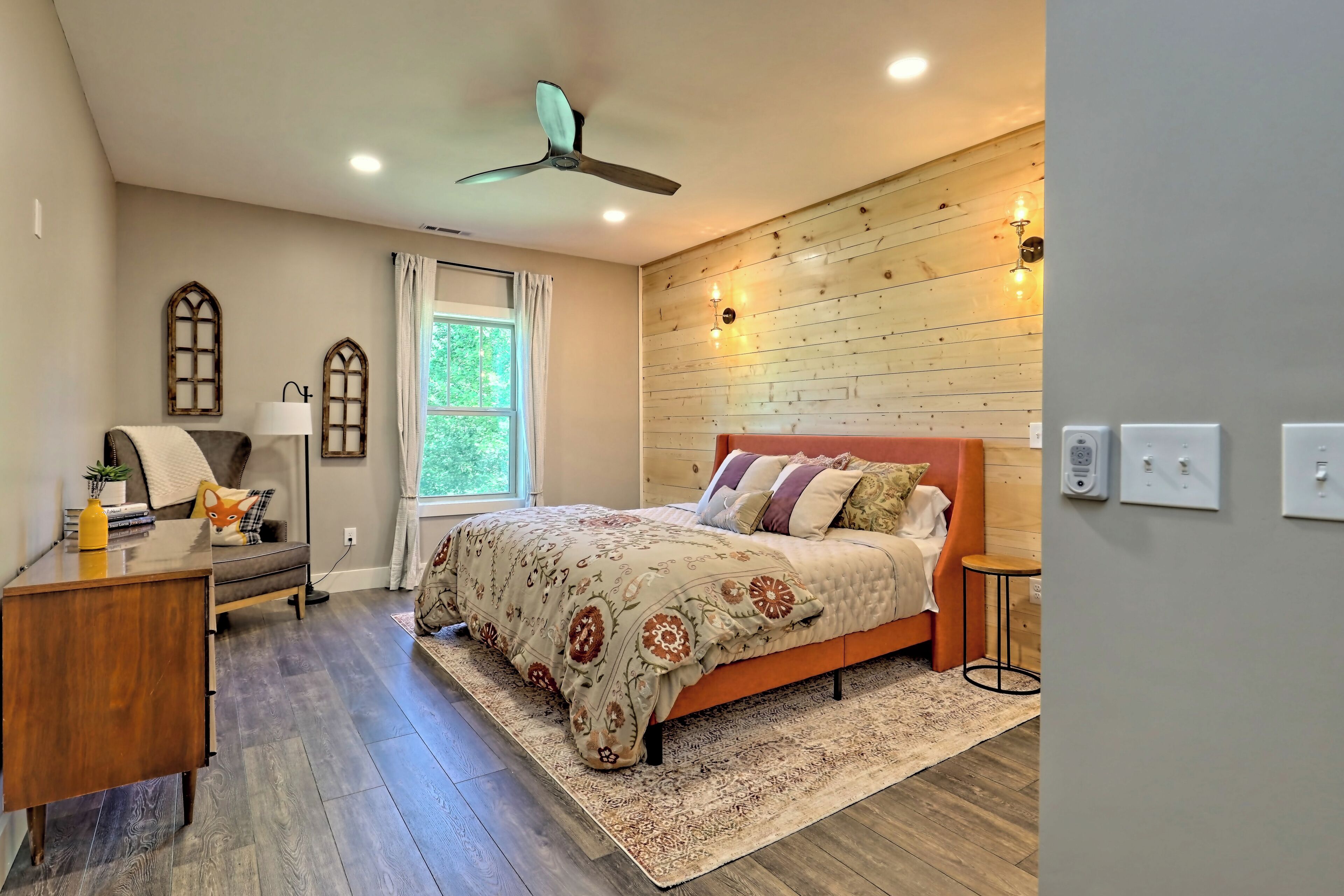 A bedroom with a wooden headboard, a bed with patterned bedding, a desk with a lamp, and a window with curtains.