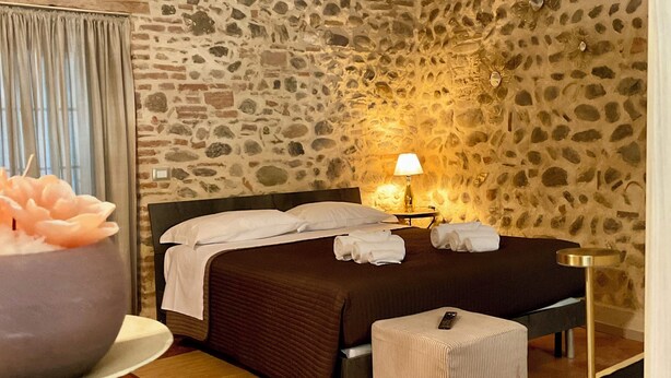 A bedroom with a stone wall, a bed with a brown bedspread, a bedside table with a lamp, and a small stool.