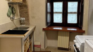 A compact kitchen with a built-in sink, a window with curtains, a radiator, and a small trash bin.