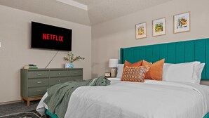 Et soveværelse med en seng, en grøn kommode, en teal sengegavl, et vægmonteret fjernsyn der viser ”NETFLIX” og tre billeder i rammer.