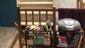Una habitación con una silla de madera, una mesa con libros y un zapato, y una estantería con cajas.