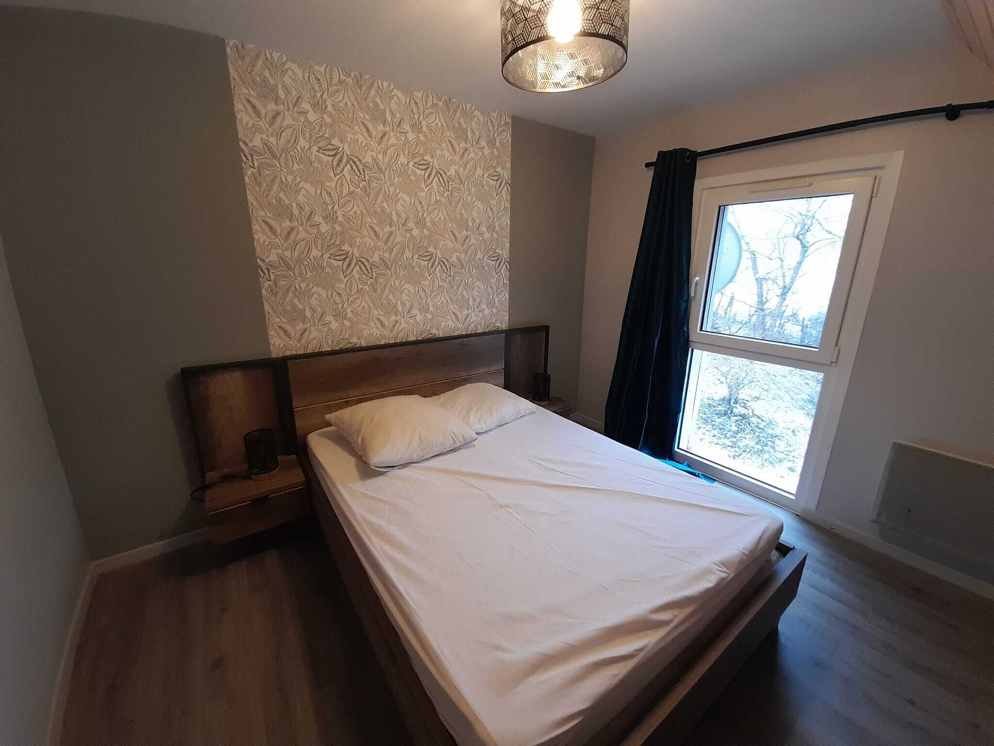 A bedroom with a wooden bed, a nightstand, a patterned wall, a window with a view of trees, and a hanging light fixture.