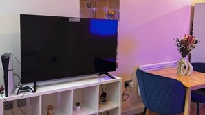 Una televisione a schermo piatto su un supporto bianco, un piccolo ripiano con oggetti decorativi e un tavolo con un vaso di fiori.