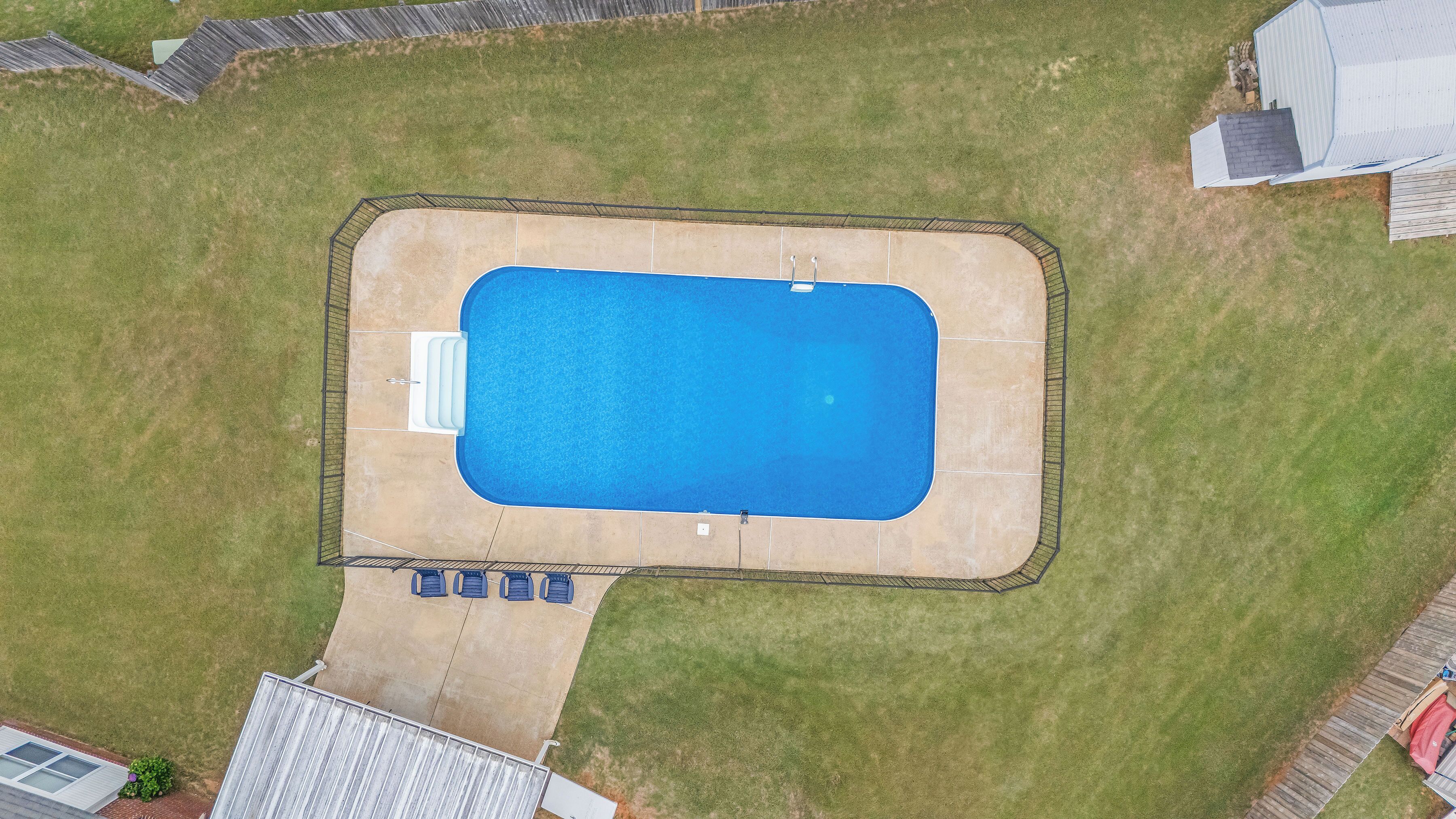 An aerial view of a rectangular swimming pool with a surrounding deck, a small set of lounge chairs, and a covered area with a red canopy.