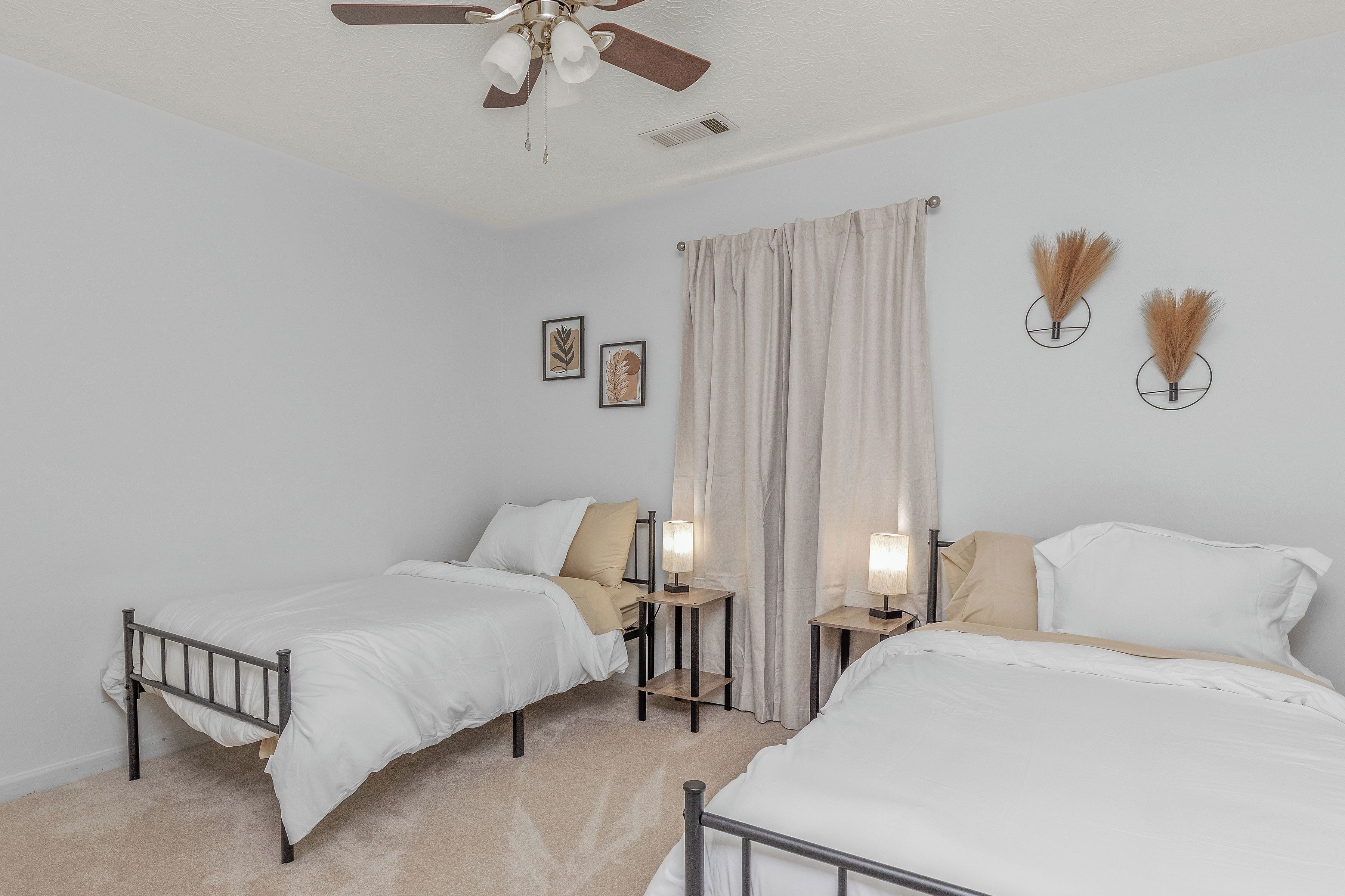 Quarto com duas camas de solteiro, ventilador de teto, abajures e janela com cortinas.
