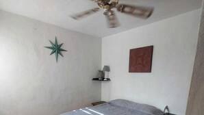 Une petite pièce propre, avec un lit, un ventilateur de plafond, une décoration en forme d’étoile, une étagère avec une lampe et un tableau accroché au mur.