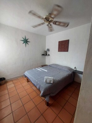 A small, clean room with a bed, a ceiling fan, a star decoration, a shelf with a lamp, and a painting on the wall.