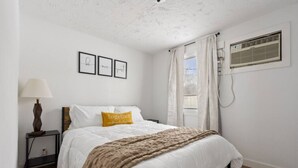 A bedroom with a bed, a nightstand, a lamp, a window with curtains, and a wall-mounted air conditioner. - Across from Leech Lake! (Walker)